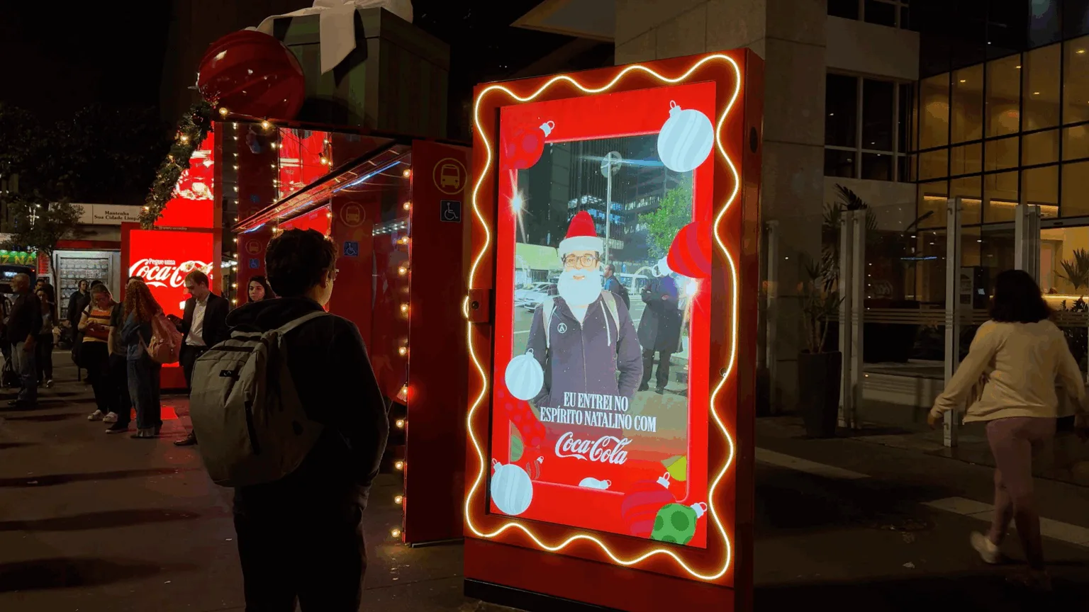 Abrigo de ônibus da Avenida Paulista decorado de Natal pela Coca-Cola, com tela digital mostrando um homem com gorro e barba de Papai Noel