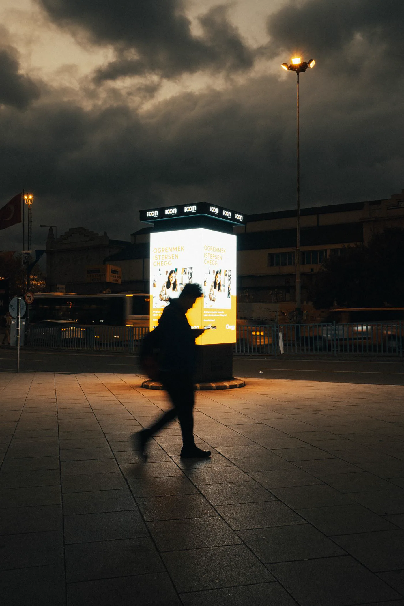 Pessoa caminhando à noite diante de um totem iluminado numa praça