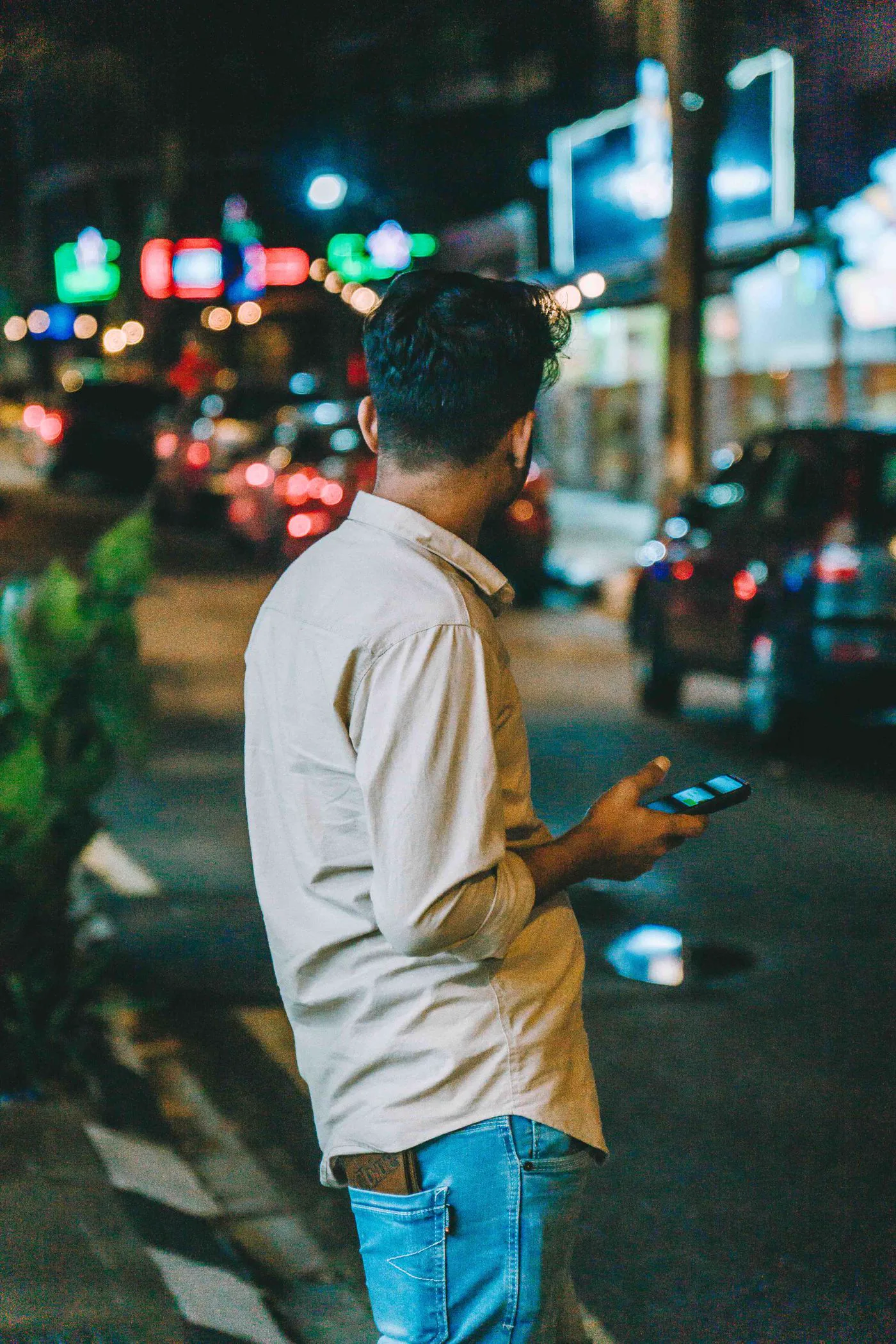 Homem mexendo no celular numa rua movimentada à noite, com luzes da cidade ao fundo